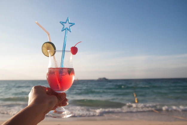 zimowy dzień marzenia o lecie, mróz na zewnątrz, palenie e-papierosa na plaży, Chorwacja lato, drink z palemką - Kobieta ręka trzyma czerwony kolor szkła letniego drinka z plaży ...